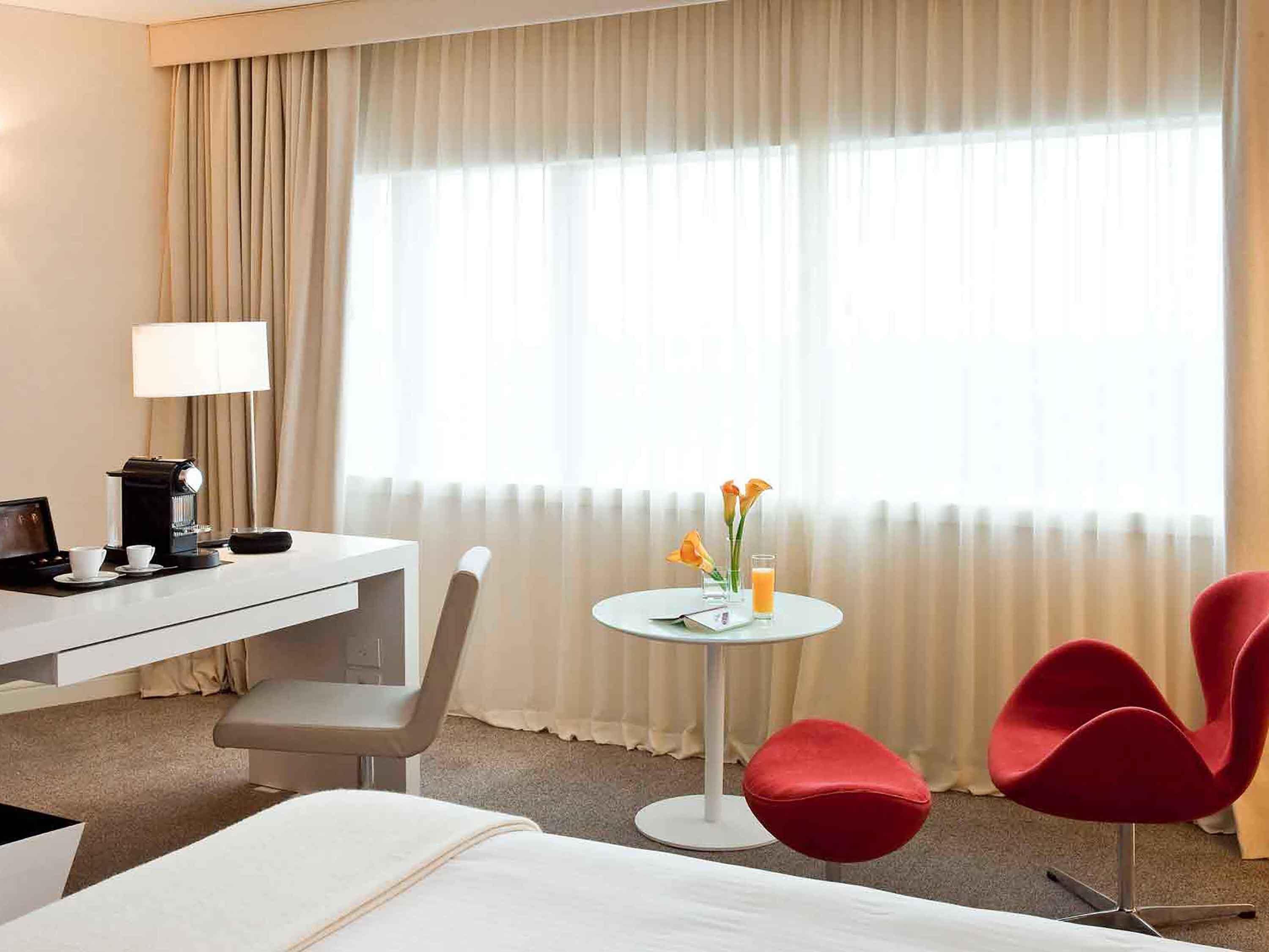 Habitación de hotel con cama, escritorio con lámpara, cafetera, pequeña mesa con vaso de zumo de naranja y flores, y dos sillas rojas.