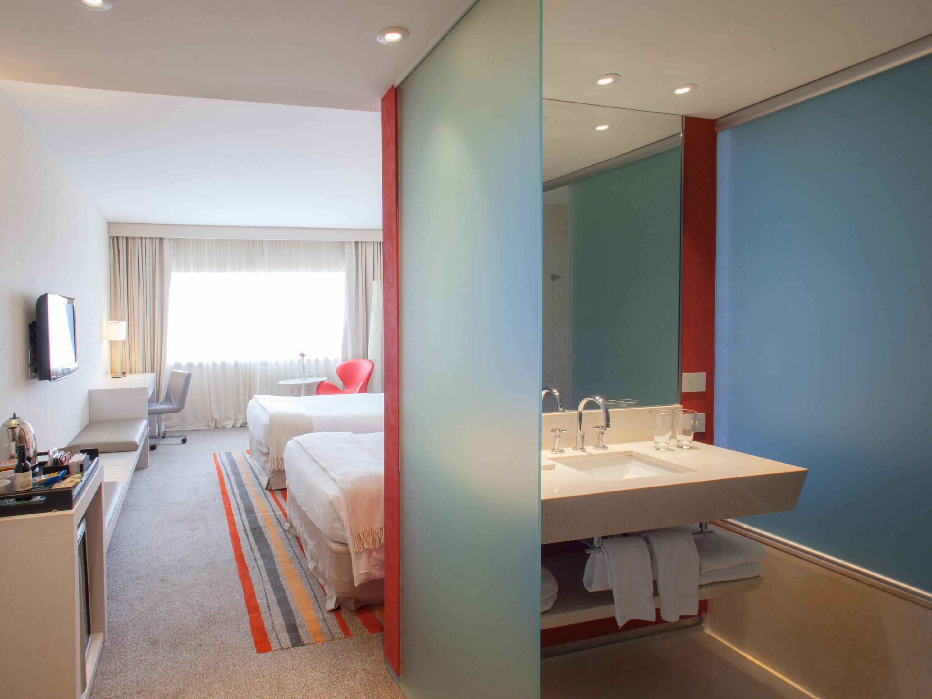 Ein Hotelzimmer mit Bett, Schreibtisch, Stuhl, Fernseher, einem Fenster mit Vorhängen sowie einem Badezimmer mit Waschbecken und Spiegel.