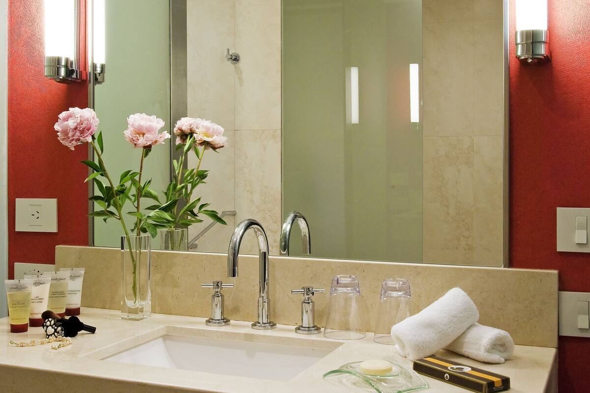 Un baño con un espejo grande, un lavabo, un perchero para toallas y un florero con flores.