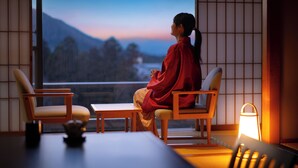 Een kamer met tatami vloerbedekking, een lage tafel, twee stoelen en uitzicht op bergen door een groot raam.