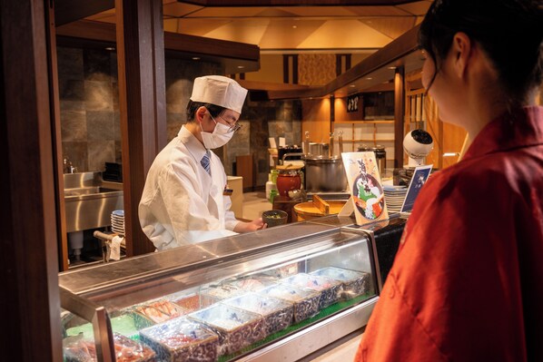 A food display case with various items, a person in a chef's uniform behind the counter, and a customer in front of the counter.