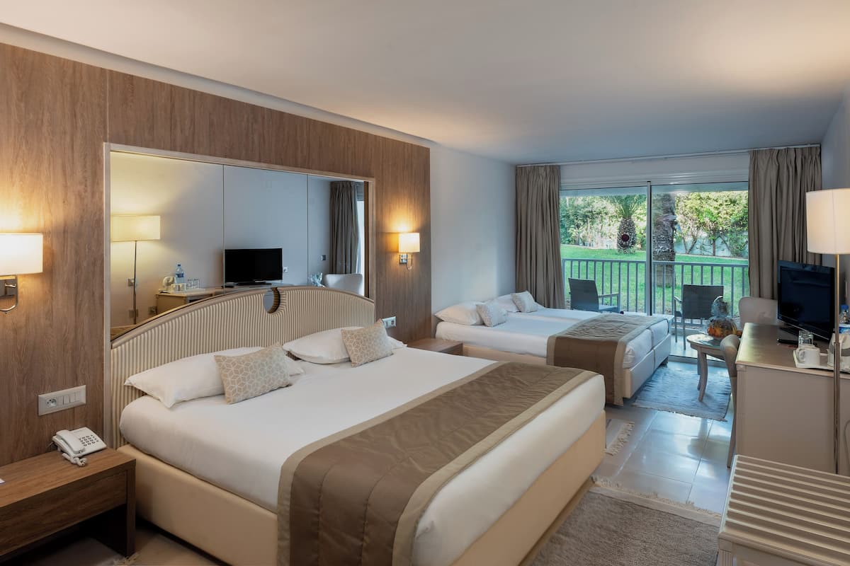 Quarto de hotel com duas camas, televisão, escrivaninha com abajur e varanda com vista.