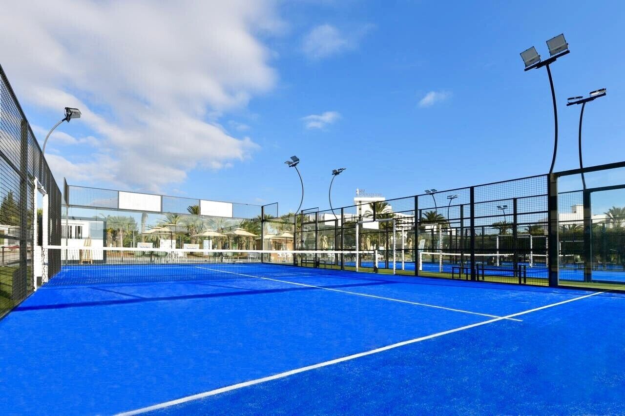 Un court de tennis intérieur doté d’une surface bleue, entouré de murs en verre et équipé de projecteurs.