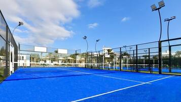 Un court de tennis intérieur doté d’une surface bleue, entouré de murs en verre et équipé de projecteurs.