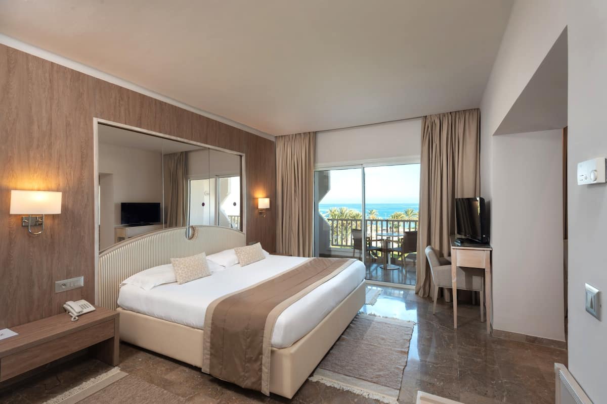 Quarto de hotel com cama, escrivaninha, televisão, varanda com vista e telefone na mesa de cabeceira.