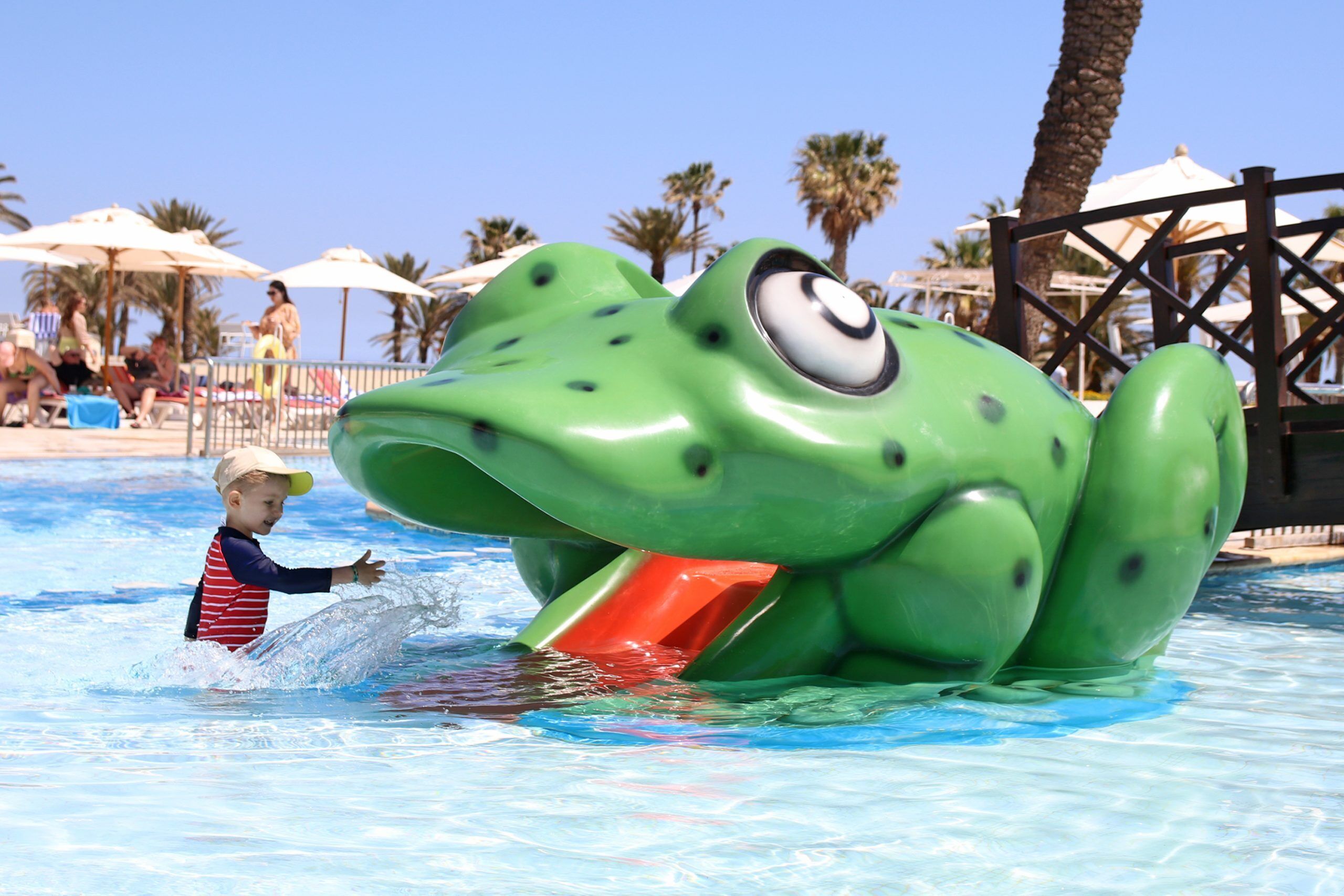 A child playing in a pool with a large frog water slide.