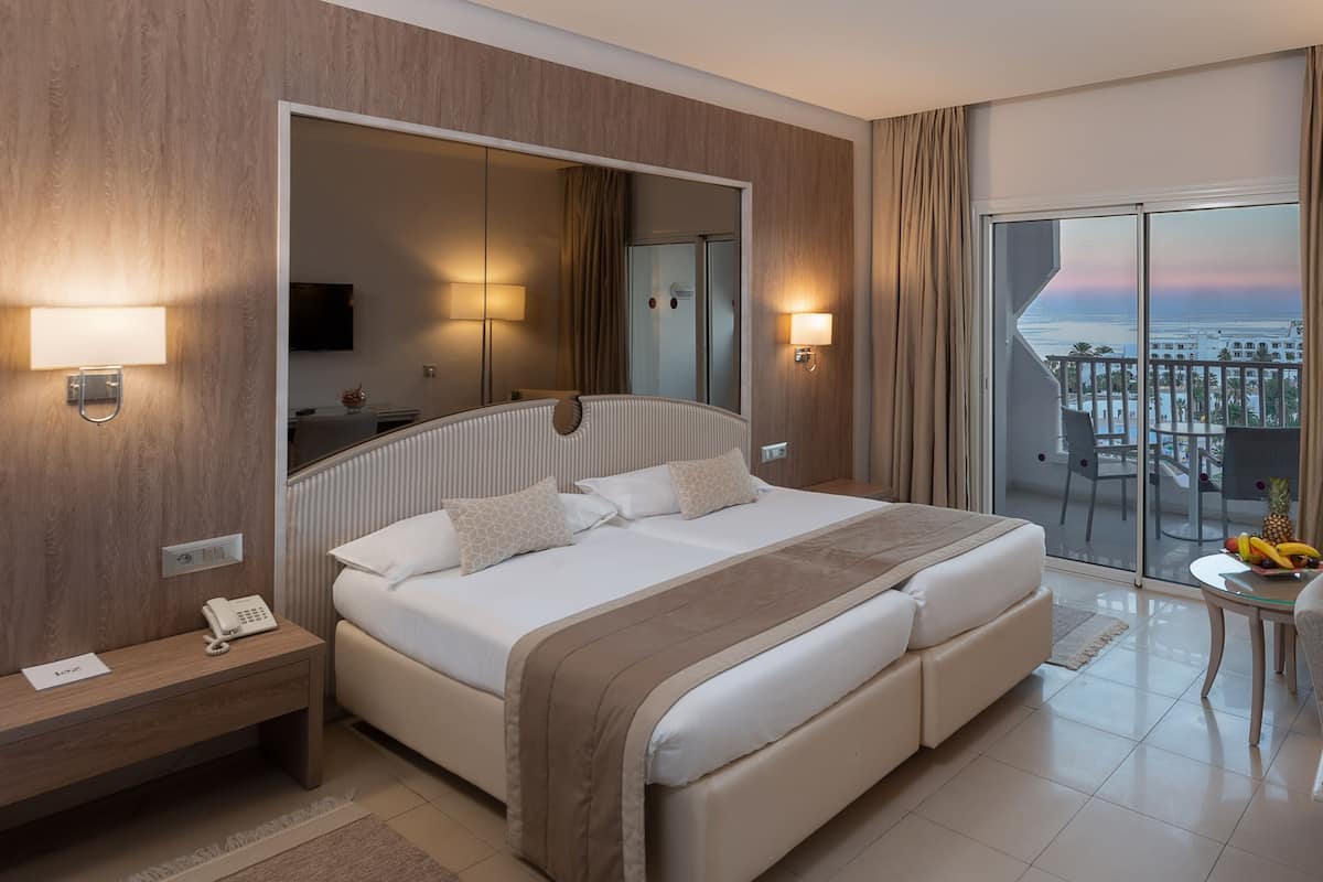 Quarto de hotel moderno com uma cama grande, abajures, um espelho e varanda com vista para a cidade.