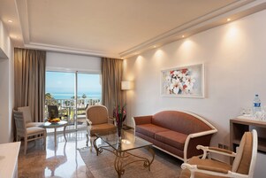 Habitación de hotel con balcón, un sofá, dos sillones, una mesa de centro de vidrio y un cuadro en la pared.