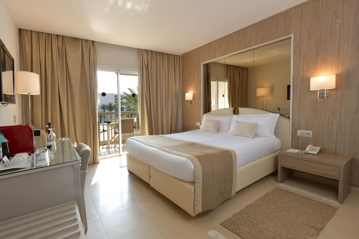 Quarto de hotel com uma cama grande, mesas de cabeceira, uma escrivaninha com luminária e vista para o exterior.
