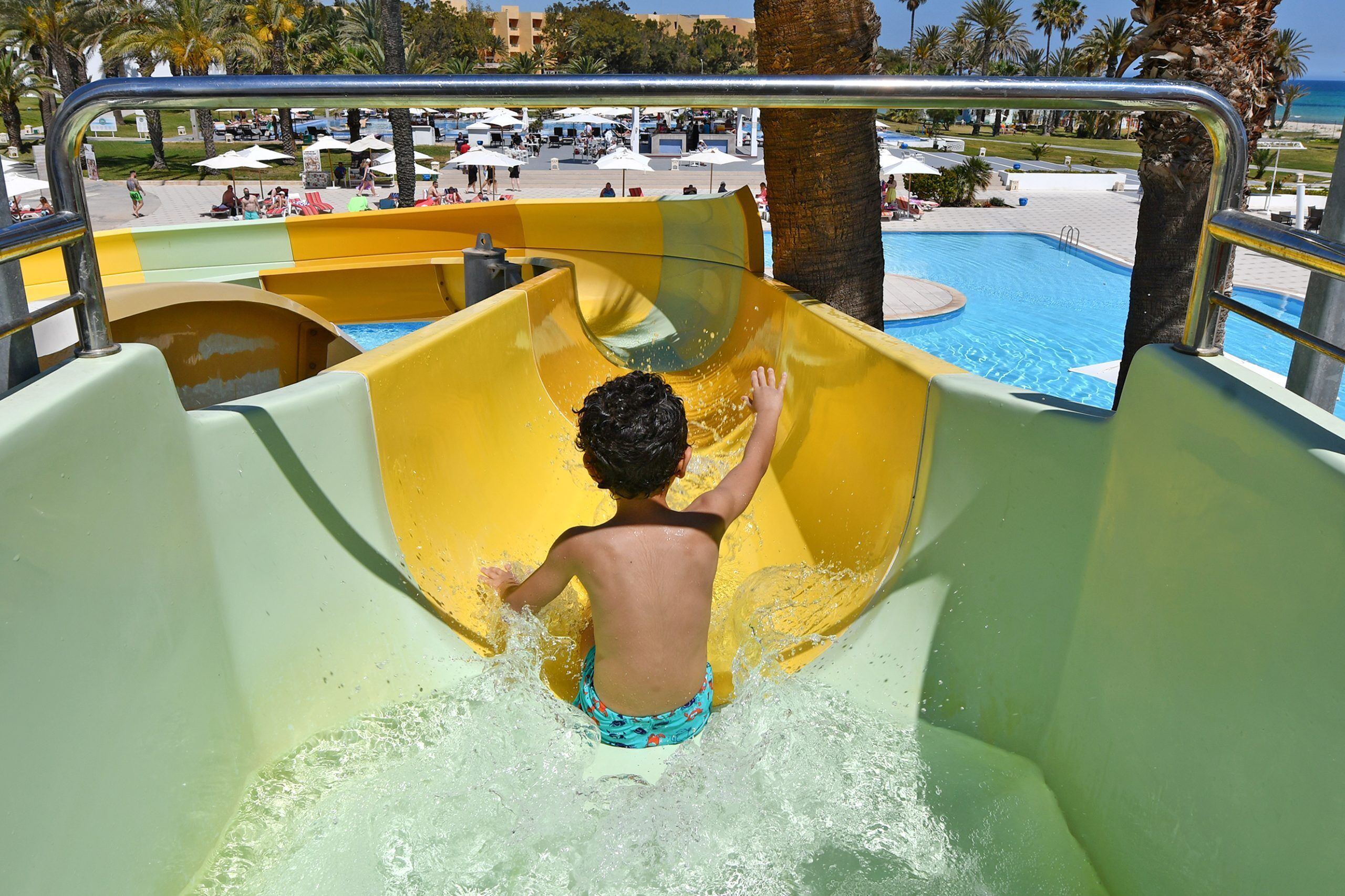 A child sliding down a yellow water slide at a poolside area with a view of the pool and palm trees in the background.
