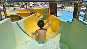 A child sliding down a yellow water slide at a poolside area with a view of the pool and palm trees in the background.