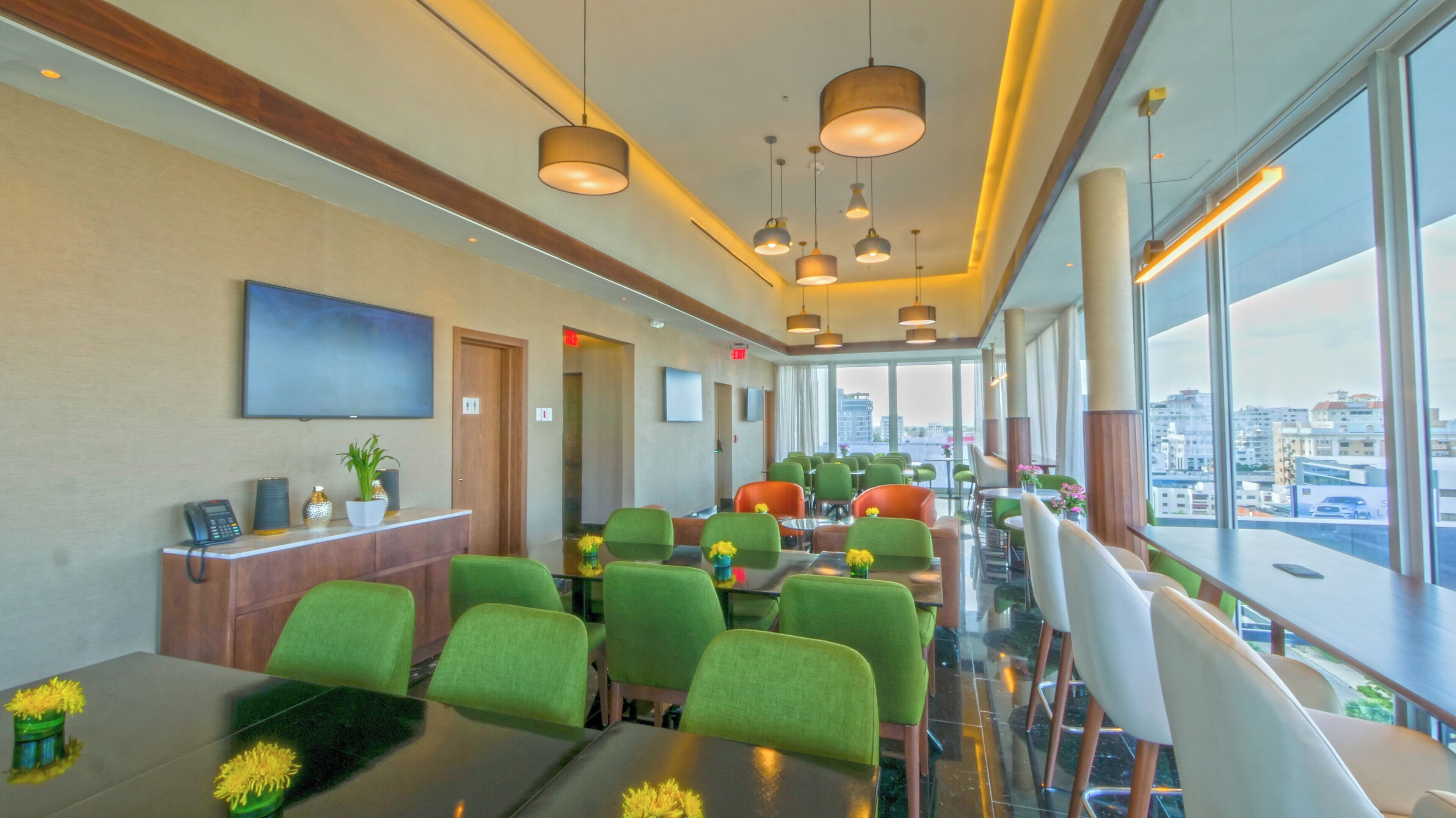 Un moderno vestíbulo de hotel con sillas verdes y naranjas, un mostrador de recepción y una pantalla de televisión grande.