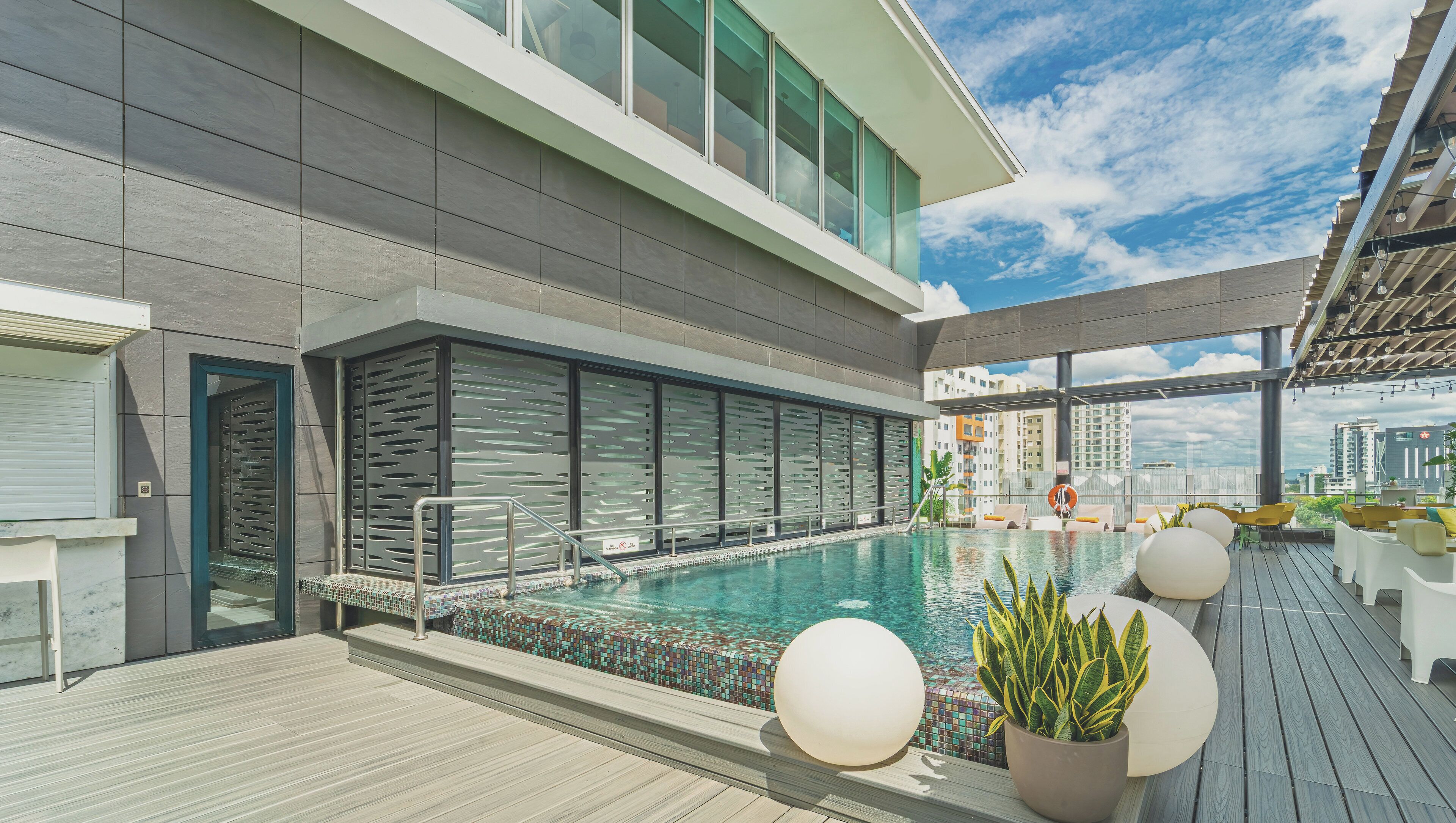 Un área de piscina en la azotea con diseño moderno, que incluye una piscina con borde de mosaicos, sillones de descanso y vistas a la ciudad.