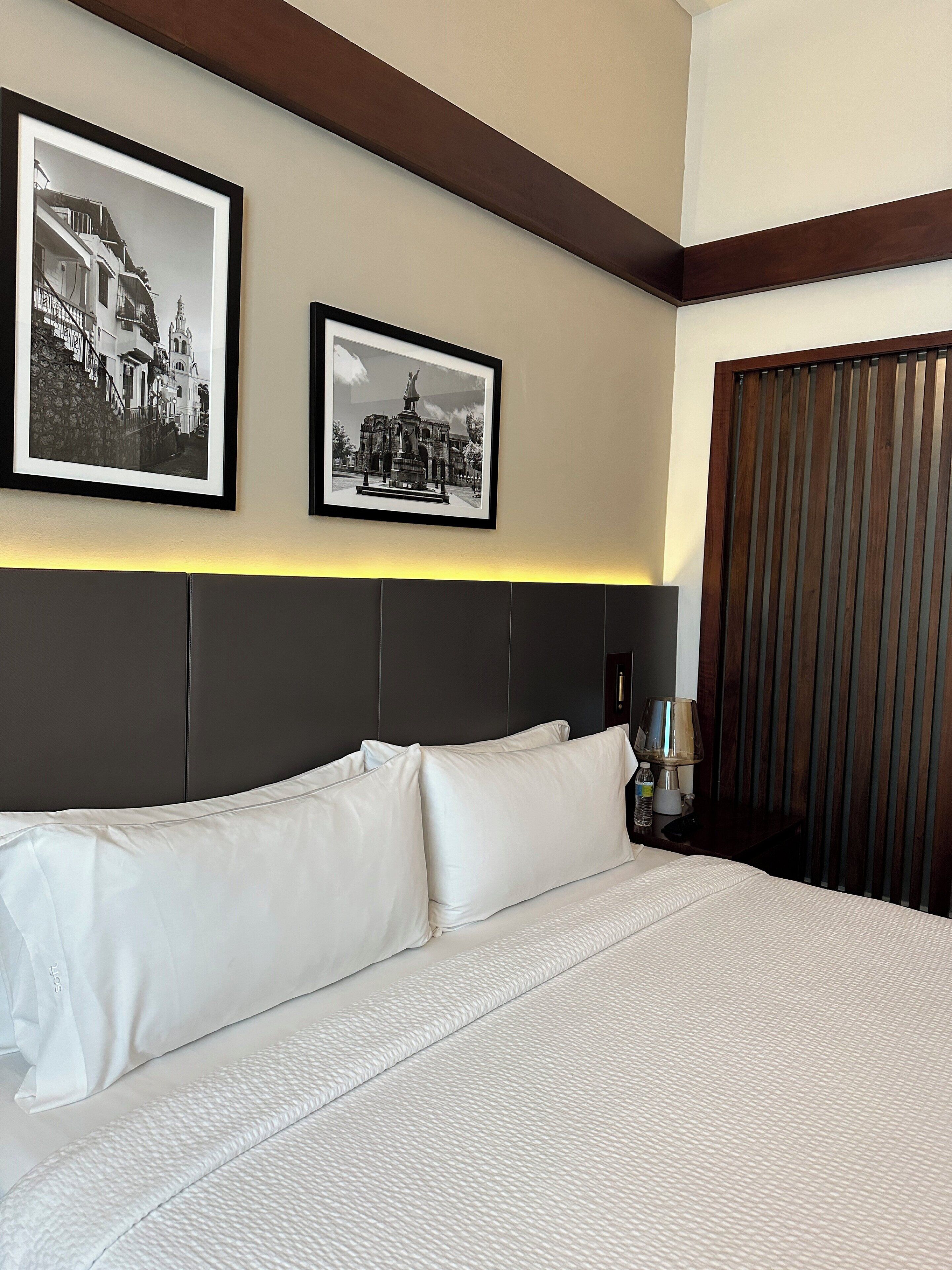 Habitación de hotel con una cama grande, dos fotografías en blanco y negro enmarcadas en la pared, un cabecero de madera y una mesita de noche con una lámpara.