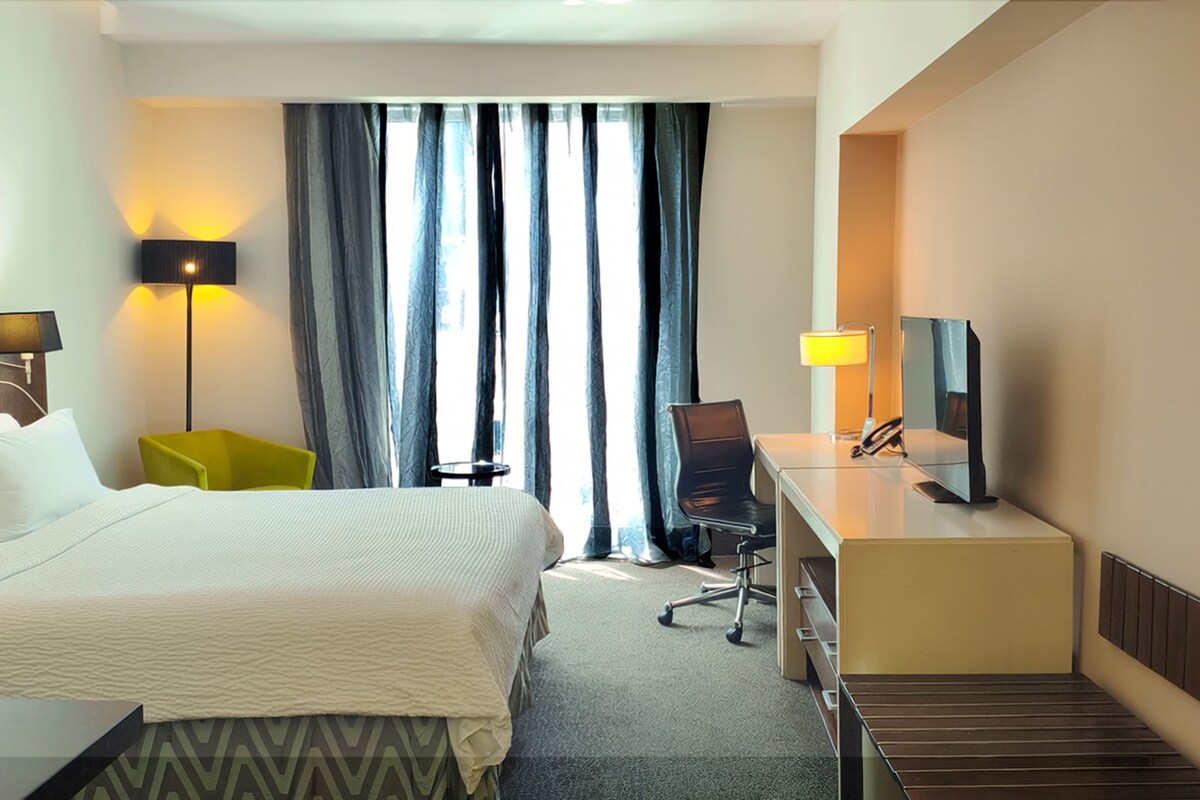 Une chambre d’hôtel équipée d’un lit, d’un bureau, d’une chaise, d’une lampe, d’une télévision et d’une fenêtre avec des rideaux.