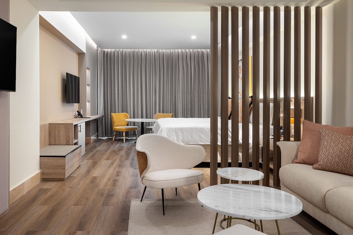 A modern hotel room with a flat-screen TV, a wooden desk, a round dining table, a sofa, and a bed with a curtain backdrop.