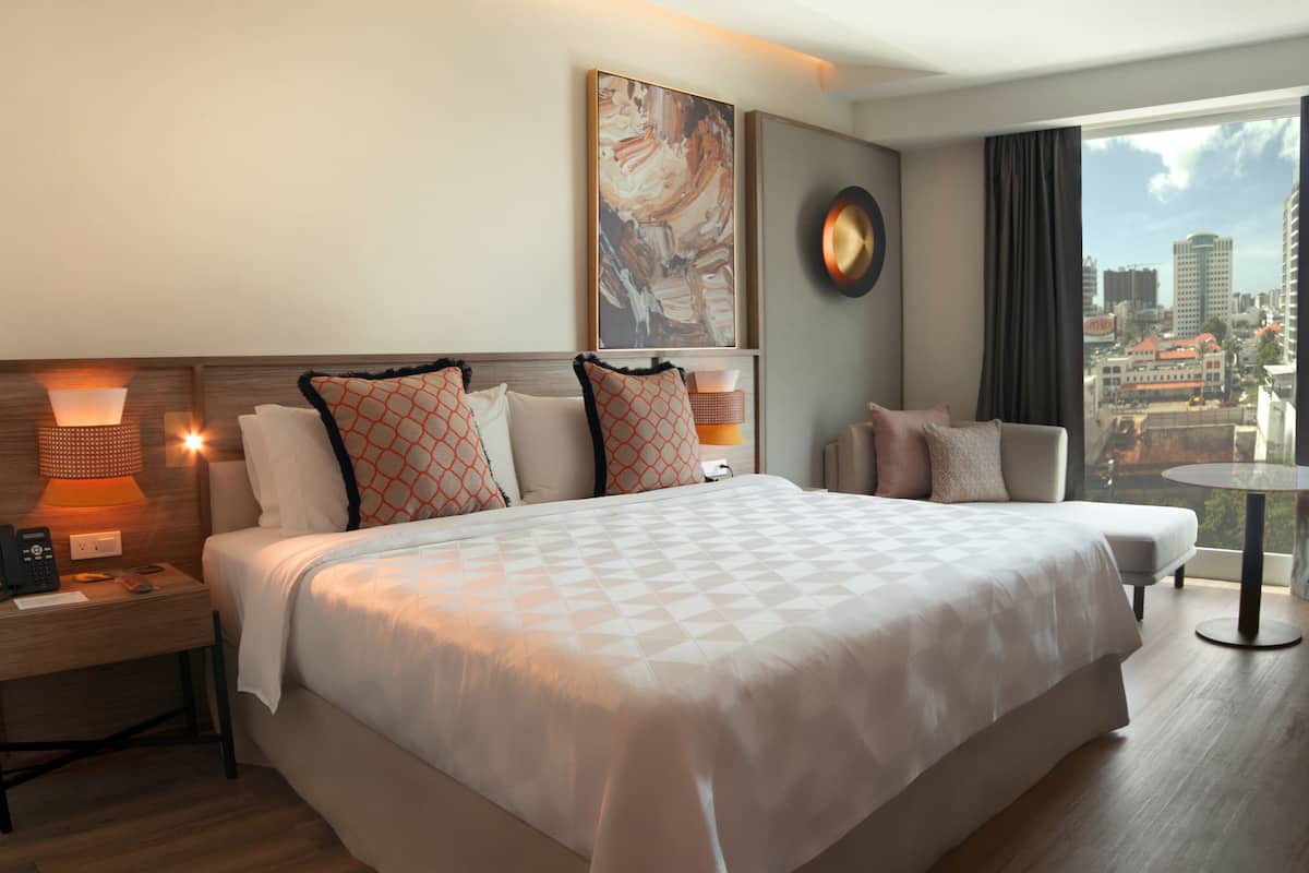 A hotel room with a large bed, a bedside table with a lamp, a clock on the wall, and a view of the city through the window.
