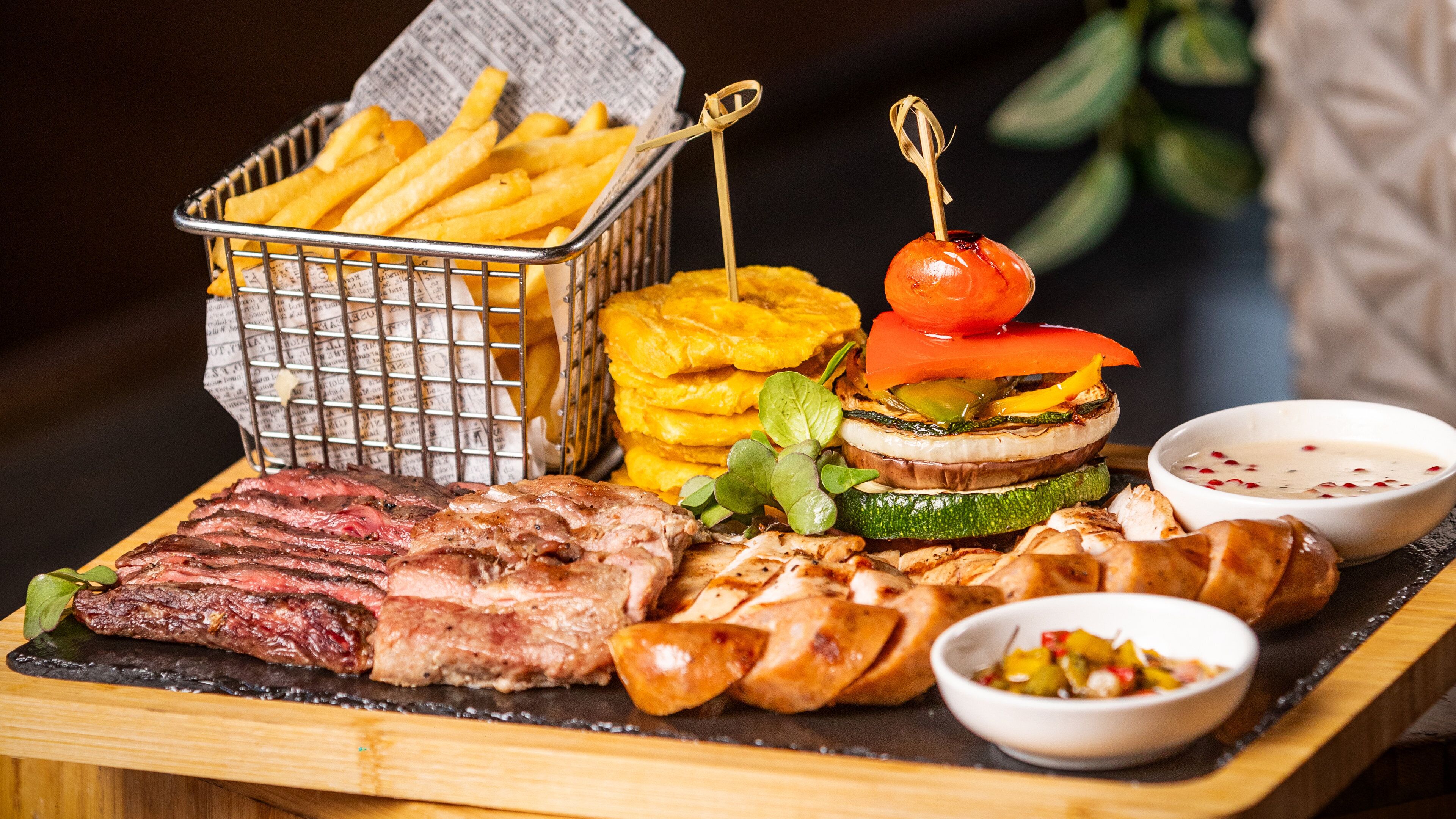 A wooden serving board with grilled meats, fried potatoes, grilled vegetables, and two bowls of dipping sauces.