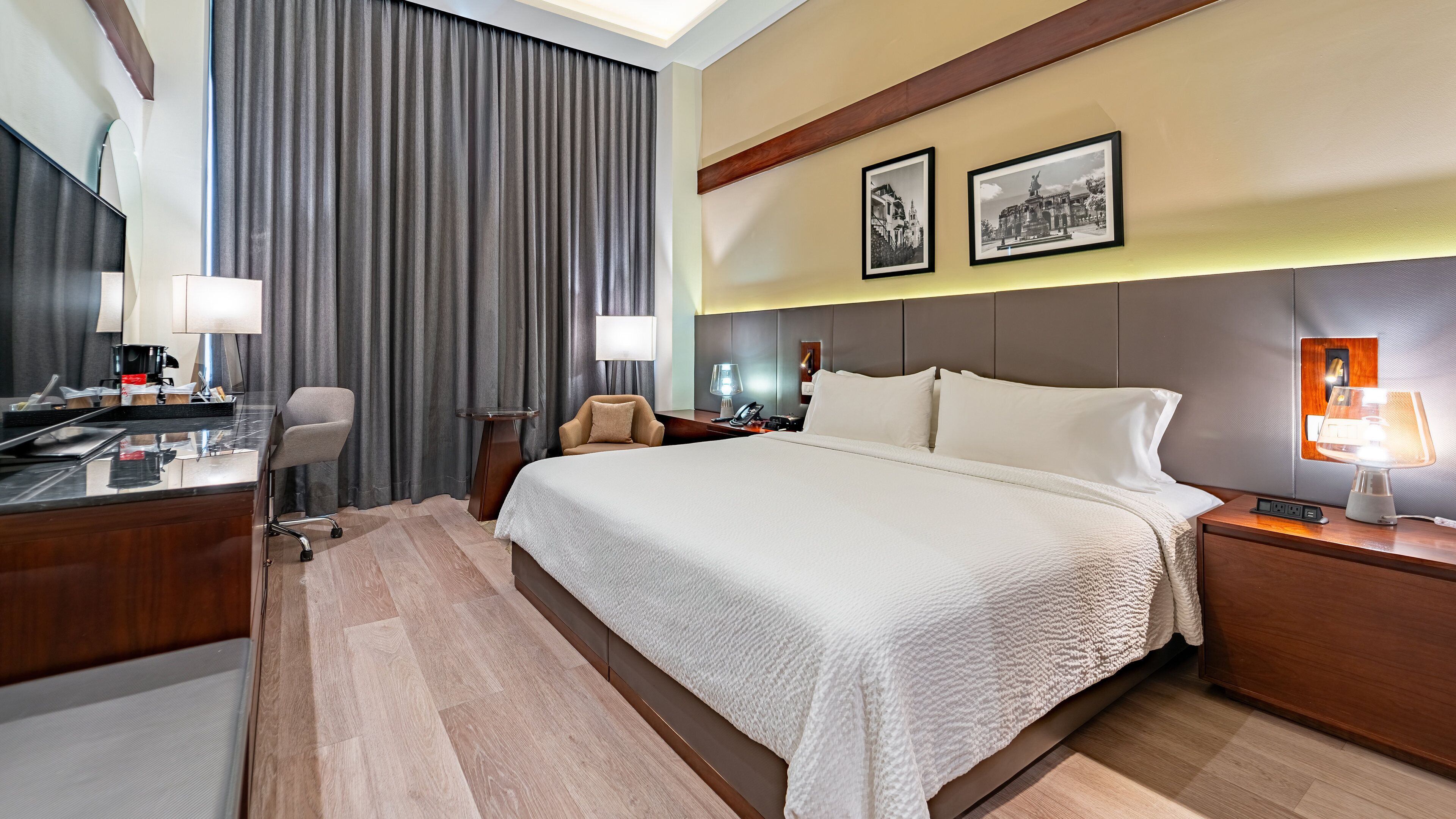 Habitación de hotel con una cama grande, un escritorio con lámpara, una silla y una mesita de noche con lámpara. En la pared hay dos cuadros enmarcados.