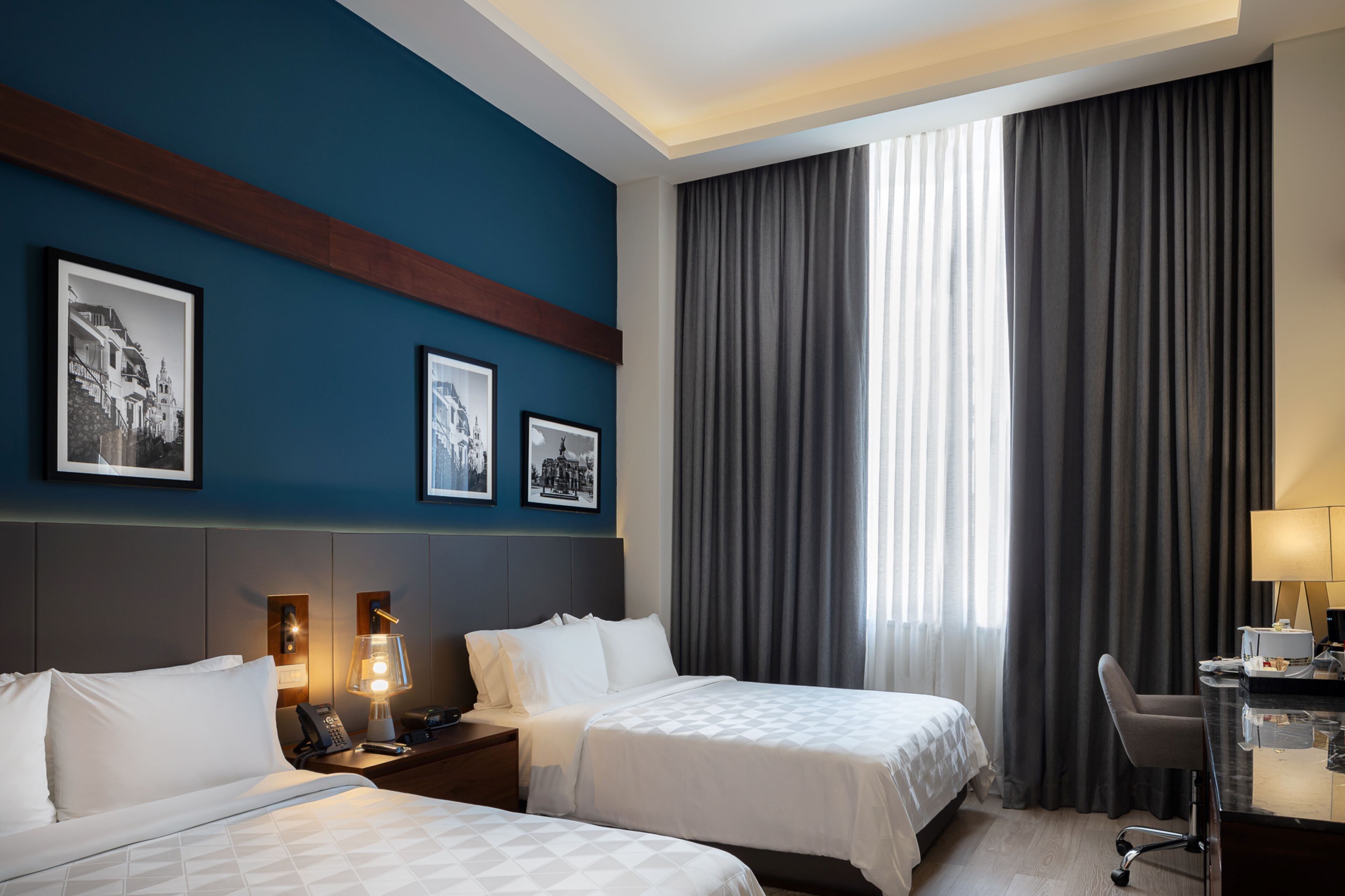 Une chambre d’hôtel avec deux lits, un bureau, une chaise et une grande fenêtre avec des rideaux.
