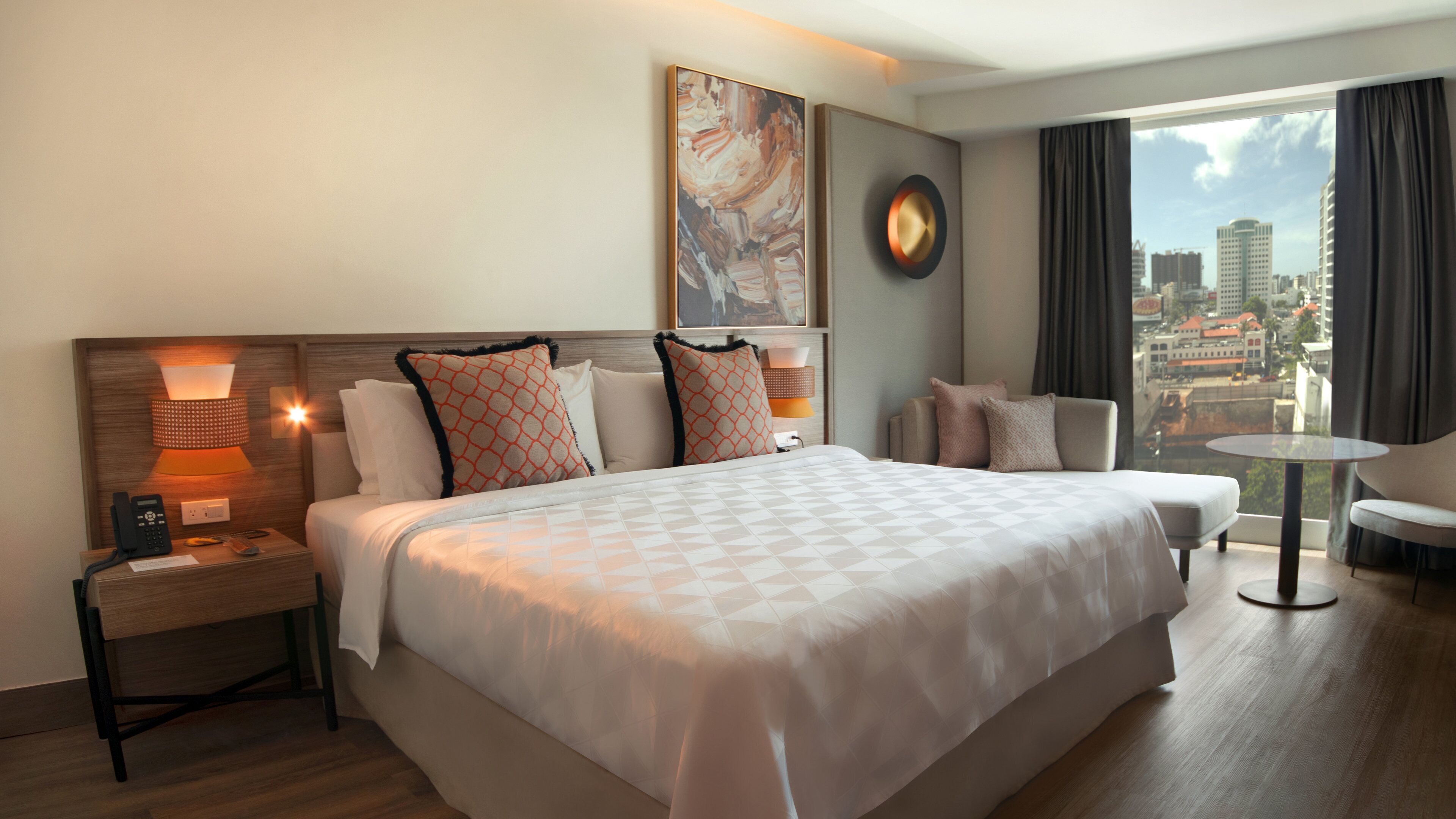 A hotel room with a large bed, bedside lamps, a clock, a city view, and a small table.