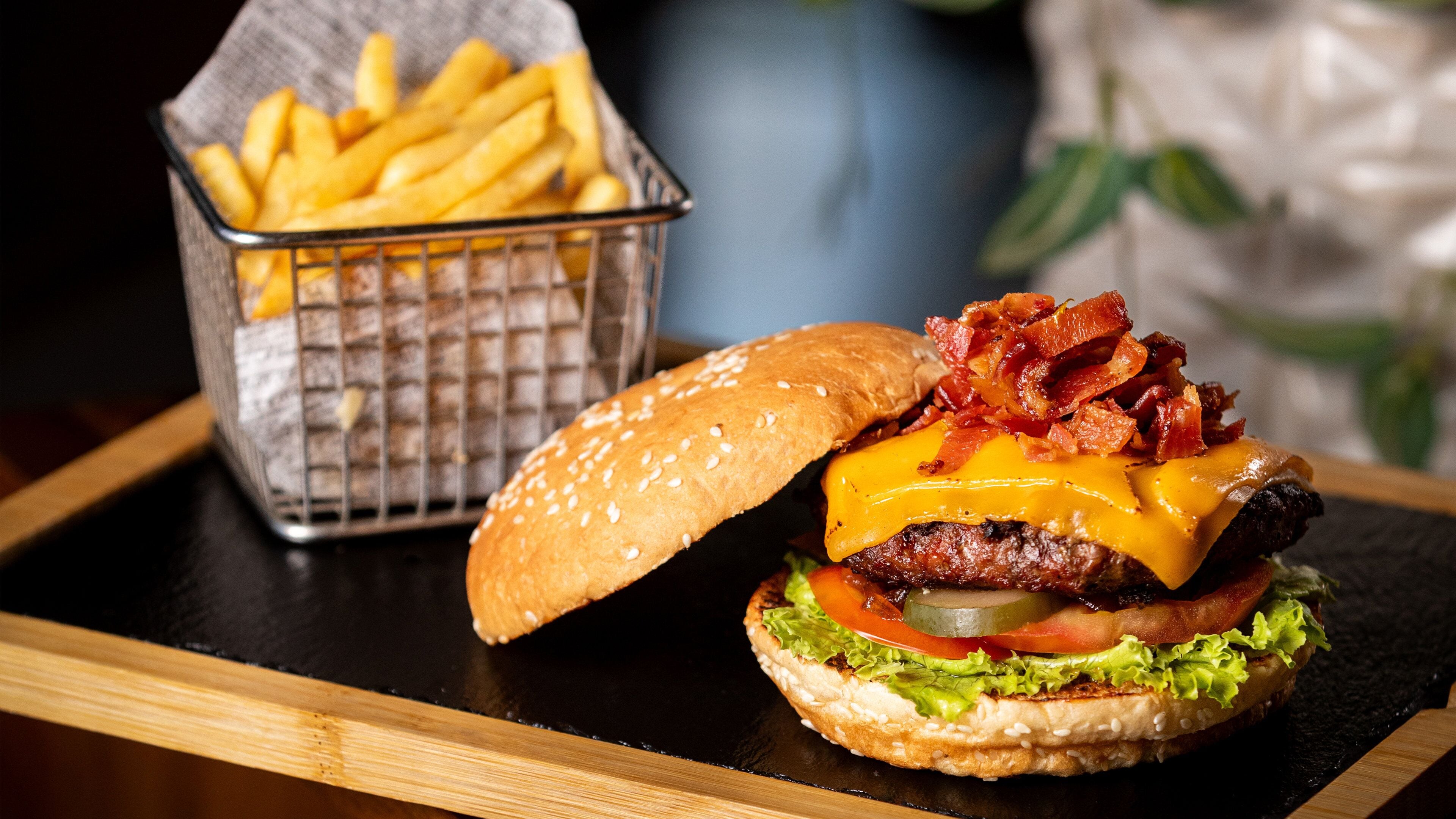 A burger with bacon, cheese, and lettuce on a bun, accompanied by a basket of fries.