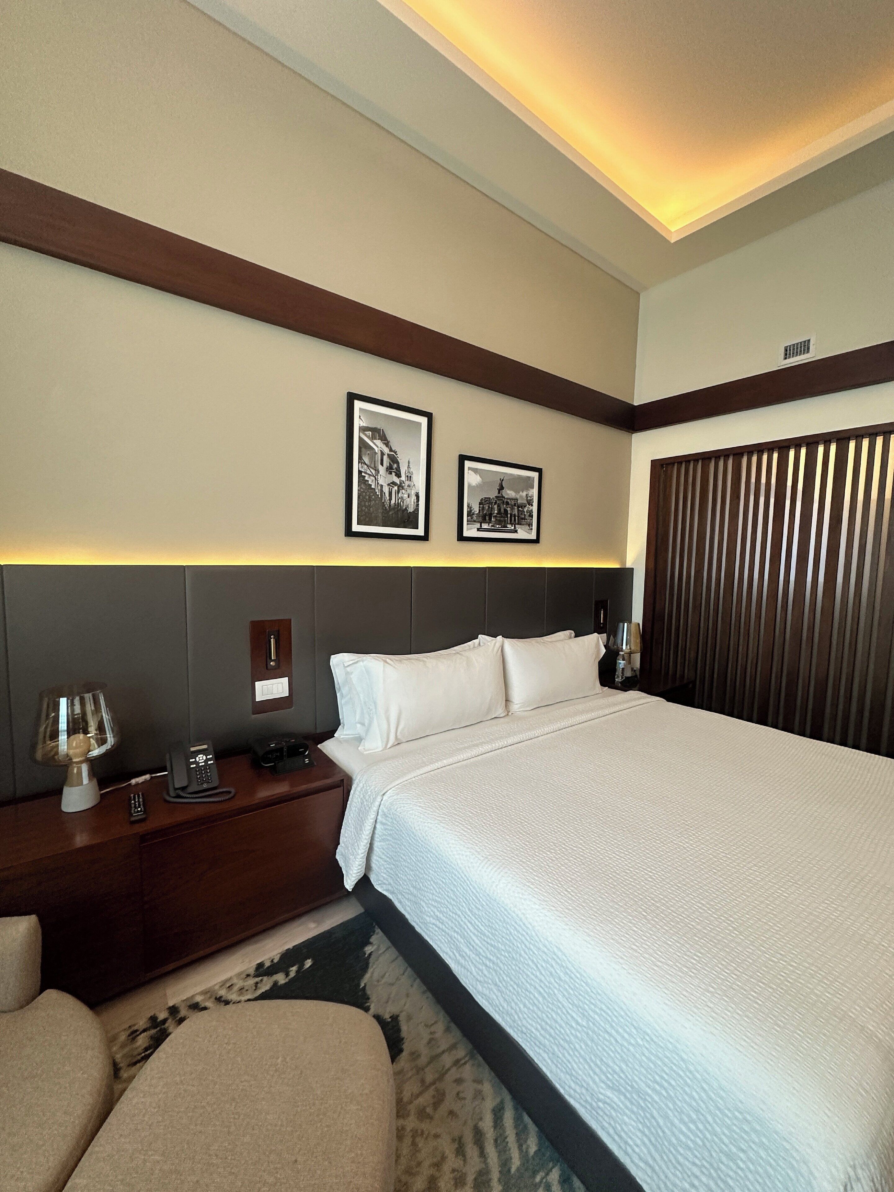 Una habitación de hotel con una cama grande, una mesita de noche con lámpara, un teléfono y dos cuadros en la pared.