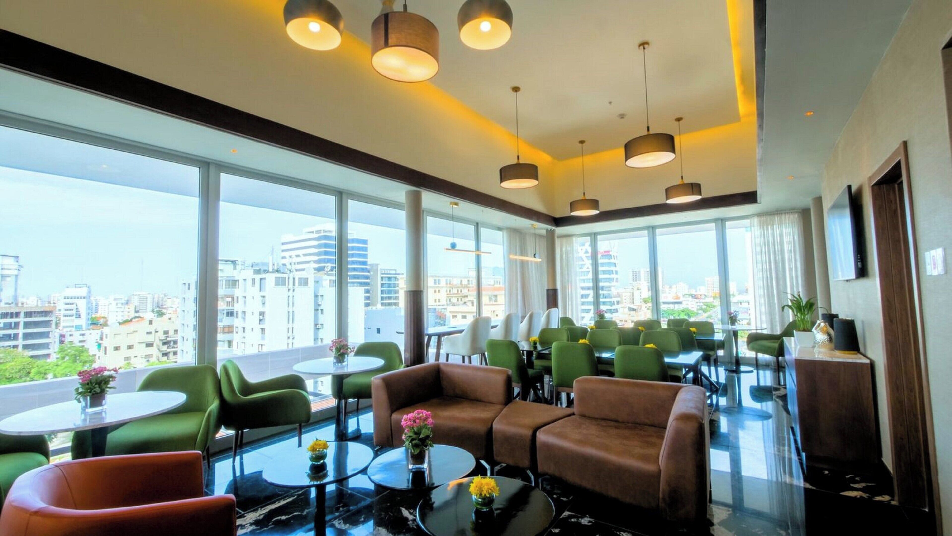 Une chambre d’hôtel moderne avec de grandes fenêtres, des meubles verts et bruns, et une vue sur la ville.