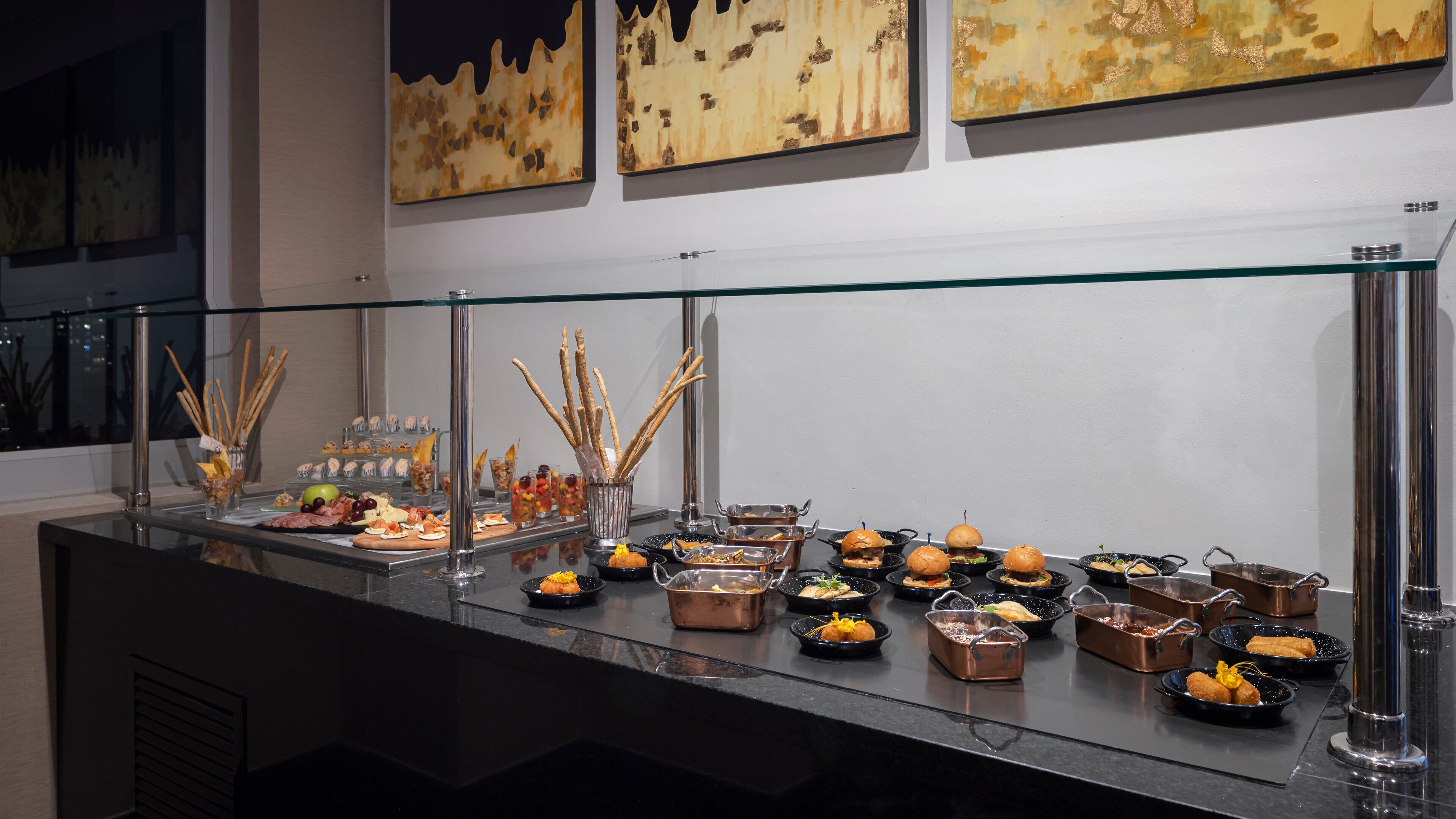 A buffet with various food items and drinks, including sandwiches, fruits, and appetizers, displayed on a glass counter with a metal railing.