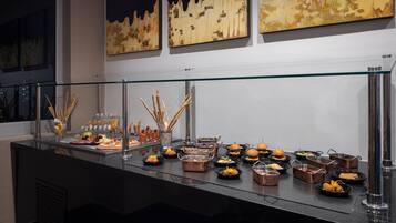 A buffet with various food items and drinks, including sandwiches, fruits, and appetizers, displayed on a glass counter with a metal railing.