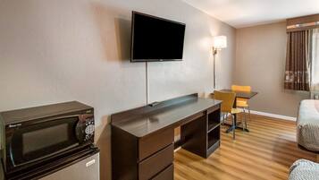Une chambre d’hôtel dotée d’un téléviseur à écran plat fixé au mur, d’une kitchenette équipée d’un four à micro-ondes et d’un réfrigérateur, d’un bureau en bois avec une chaise et d’un lit recouvert d’une couverture à motifs.