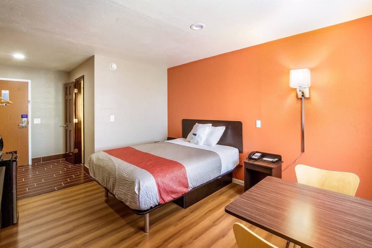 A hotel room with a bed, a desk, a chair, a nightstand, and a wall-mounted lamp.