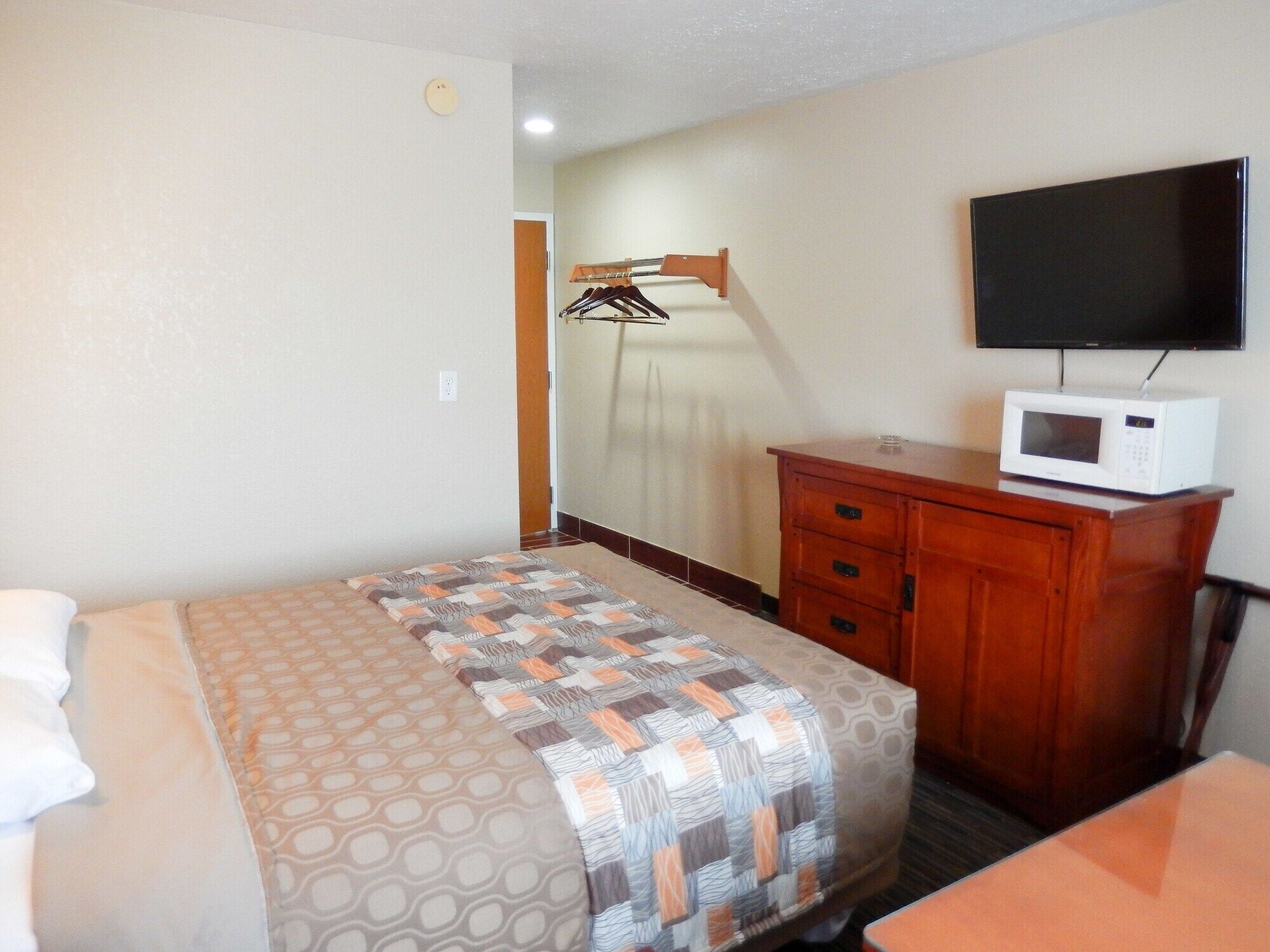 A hotel room with a bed, a wooden dresser, a microwave, and a TV on the wall.