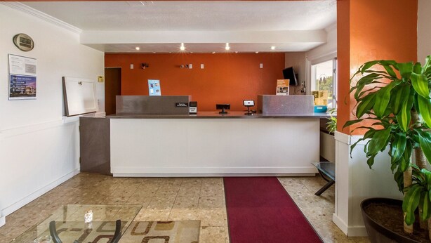 A reception area with a white reception desk, a clock, and a plant. - Motel 6 Rossford, OH (Rossford)