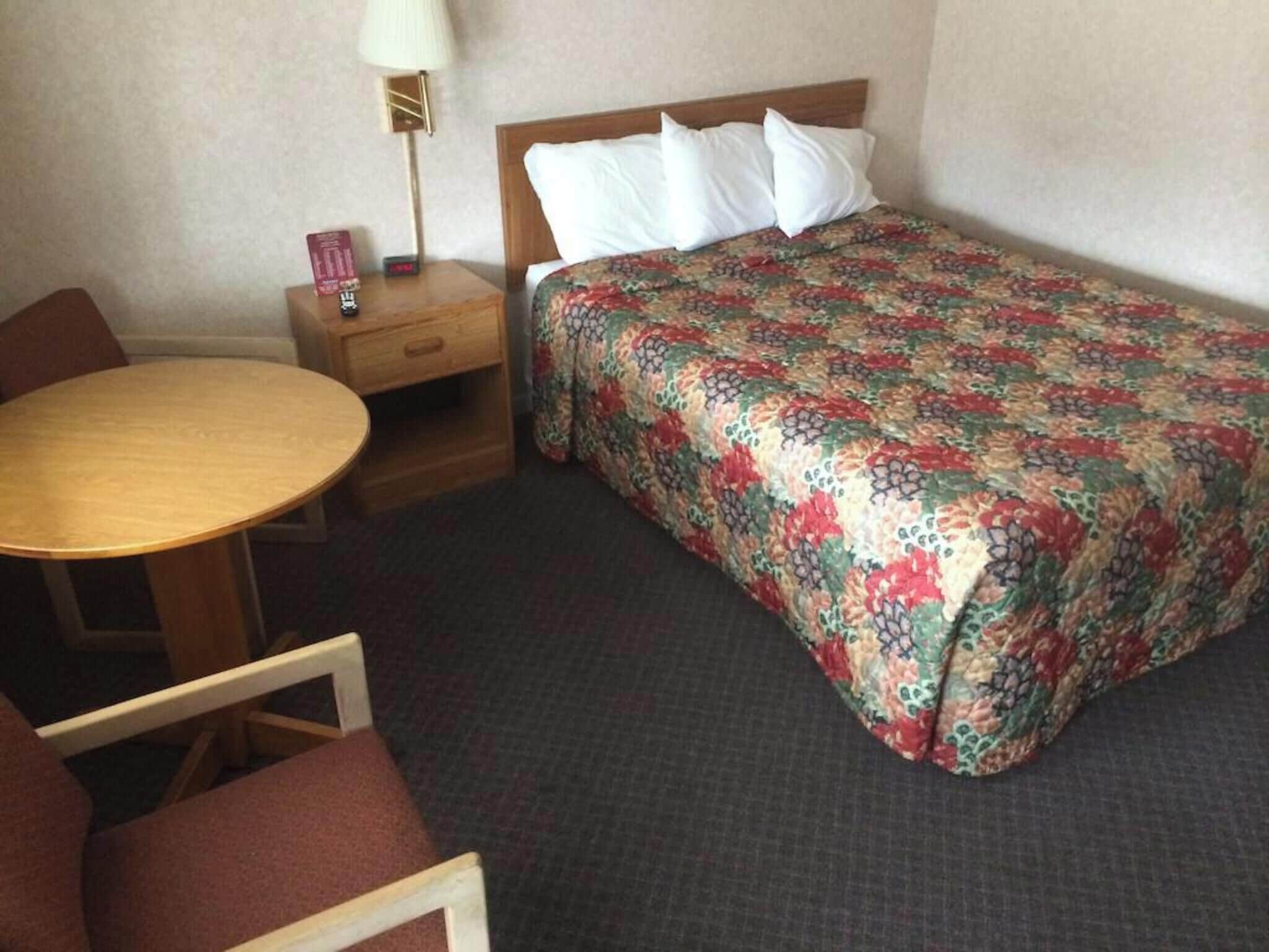 Une chambre d’hôtel avec un lit, une table de chevet en bois avec une lampe, une petite table et une chaise.