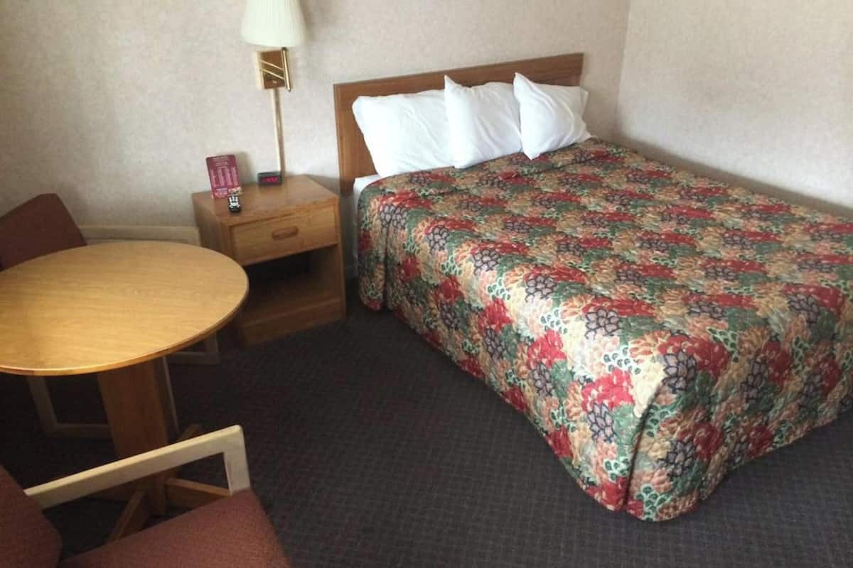 A hotel room with a bed, a wooden nightstand with a lamp, a small table, and a chair.