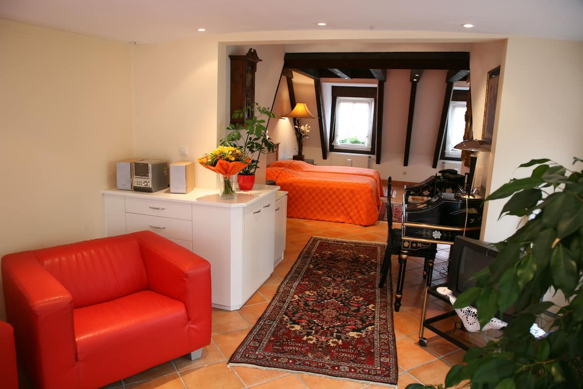 A compact room with a red sofa, a small desk with a computer and speakers, a bed with an orange bedspread, and a large window with curtains.