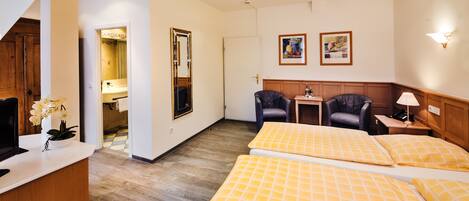 Habitación de hotel con cama, escritorio, silla, espejo y baño visible a través de una puerta abierta.