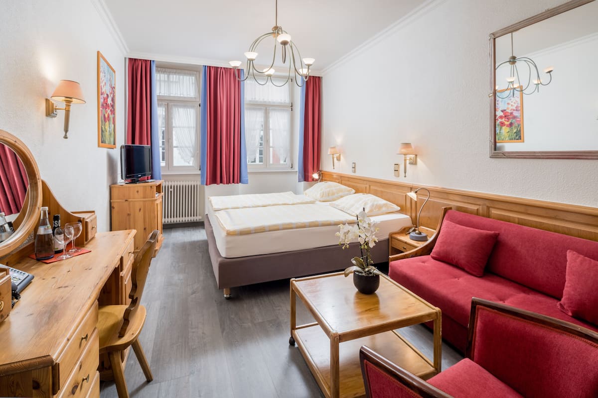 A hotel room with a double bed, a red sofa, a wooden desk with a mirror, a television, and a potted plant.