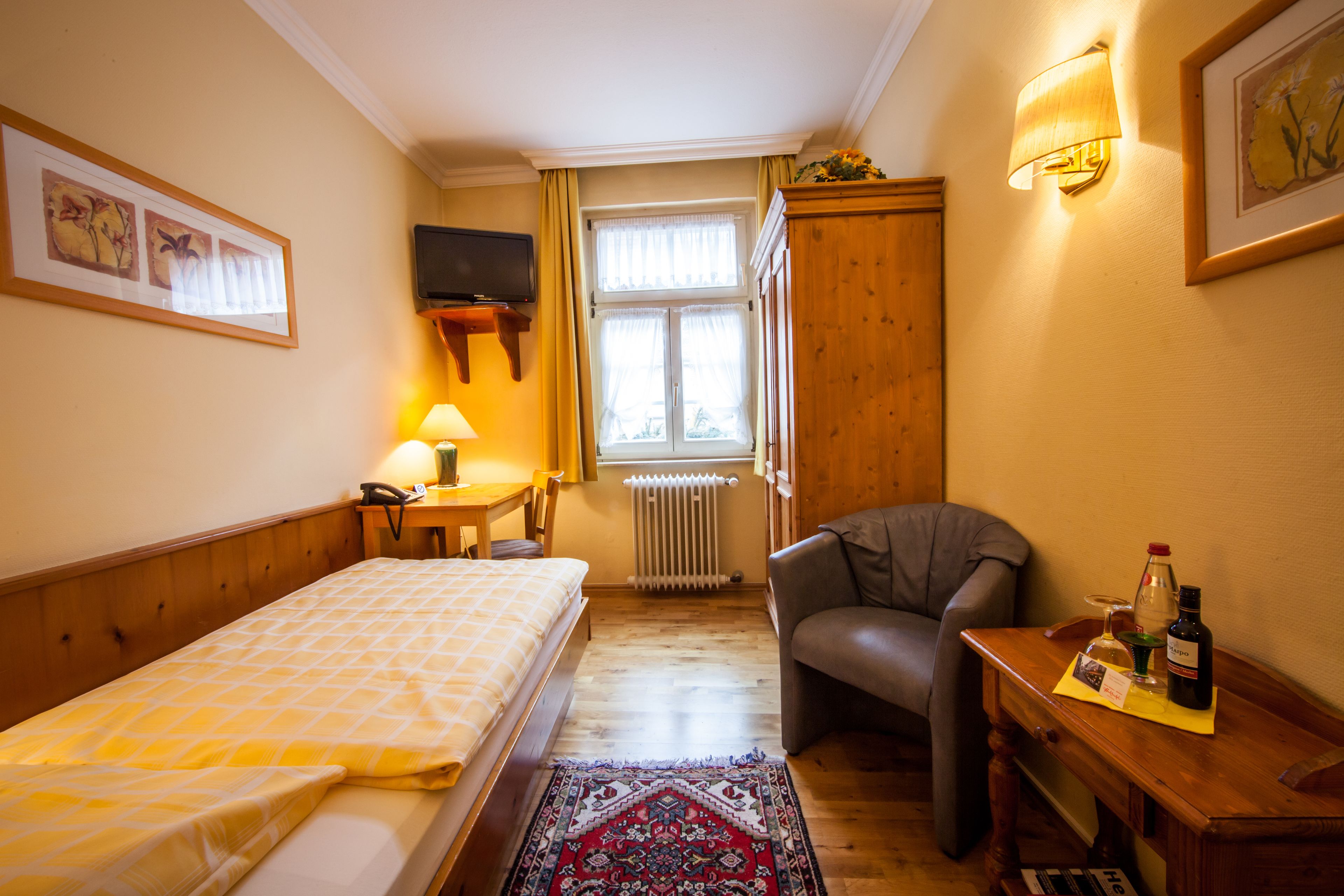 Hotellihuoneessa on sänky, tuoli, pieni pöytä, jonka päällä on pullo ja lasi, sekä hyllyllä oleva televisio.