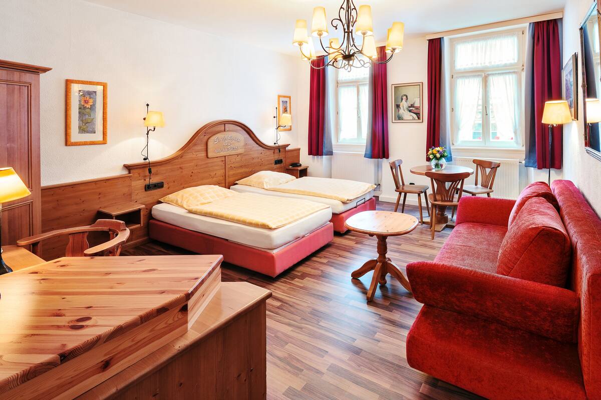 A hotel room with two beds, a wooden desk, a red sofa, a dining table, and a chandelier.