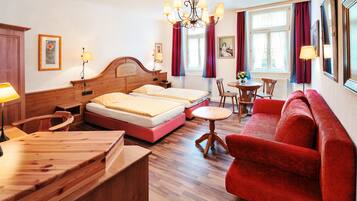 A hotel room with two beds, a wooden desk, a red sofa, a dining table, and a chandelier.