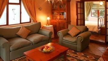 A cozy living room with a sofa, armchair, and a small table with fruit. There is a bookshelf and a window with curtains.