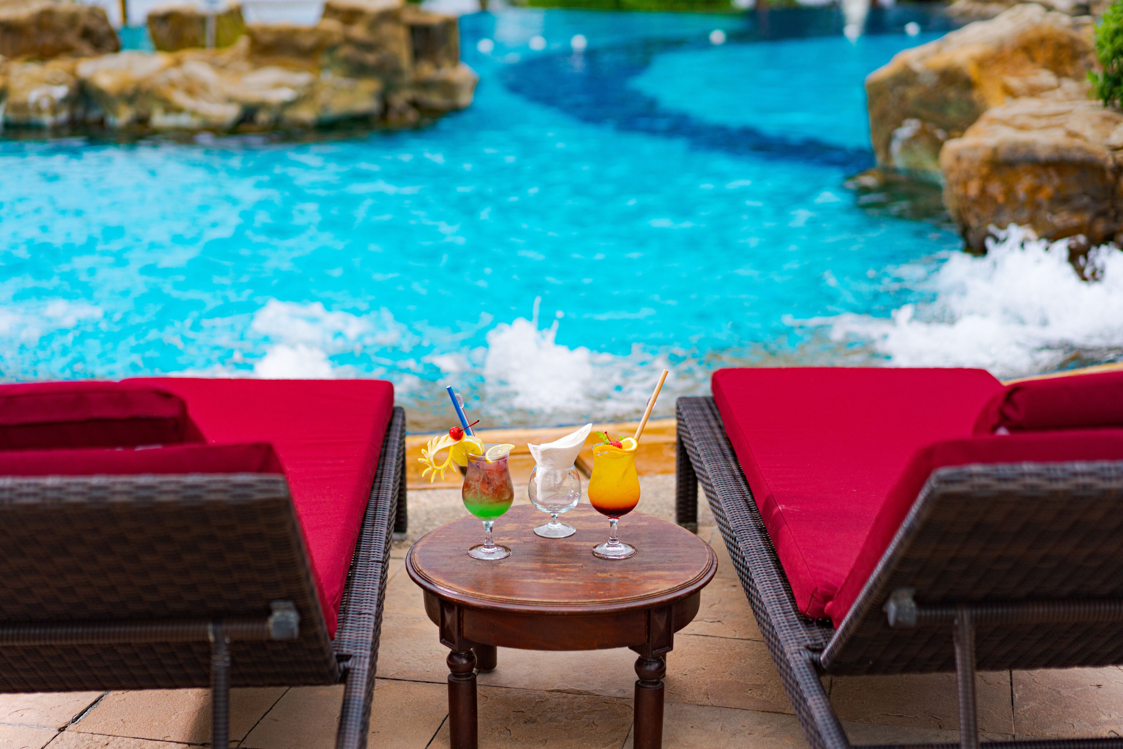 A poolside area with a table holding three drinks, two wicker lounge chairs with red cushions, and a view of a blue pool with rocks around it.