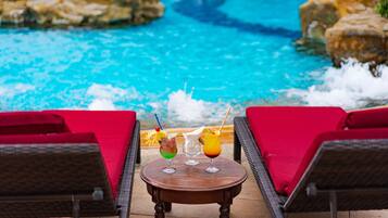 A poolside area with a table holding three drinks, two wicker lounge chairs with red cushions, and a view of a blue pool with rocks around it.