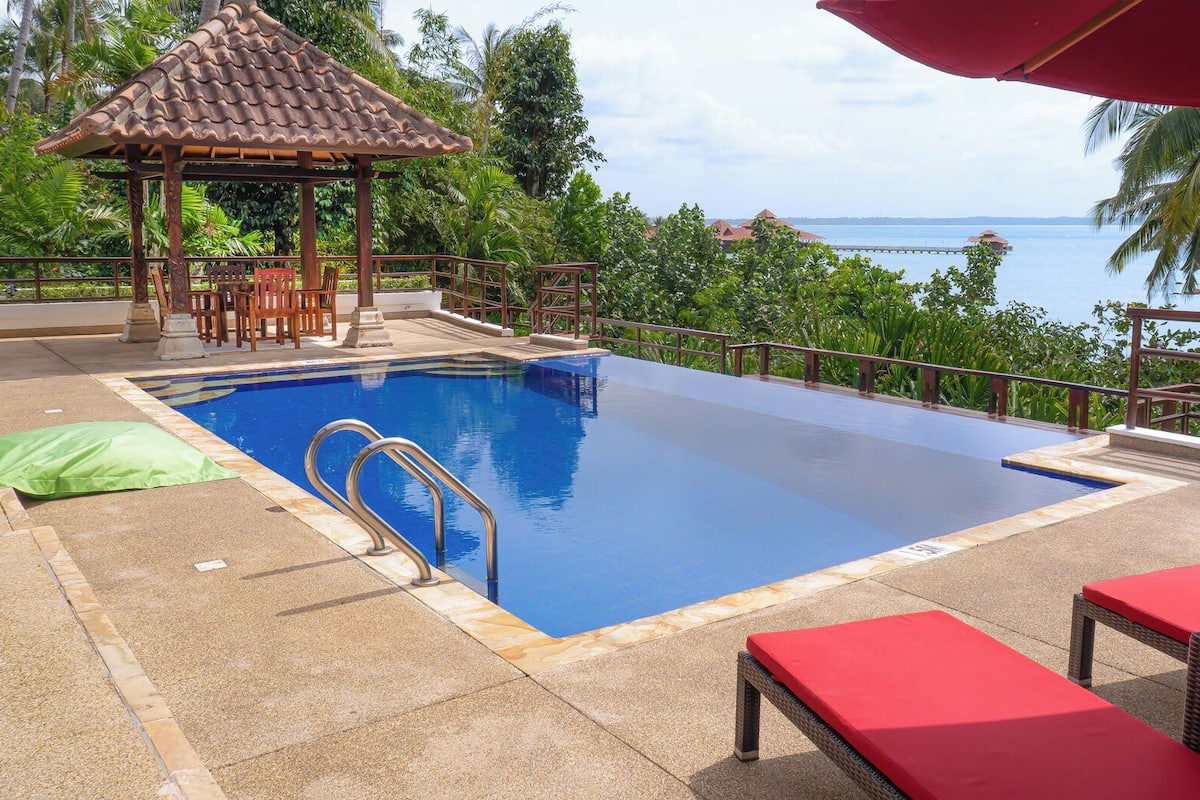 A pool with a wooden deck, lounge chairs, and a gazebo.