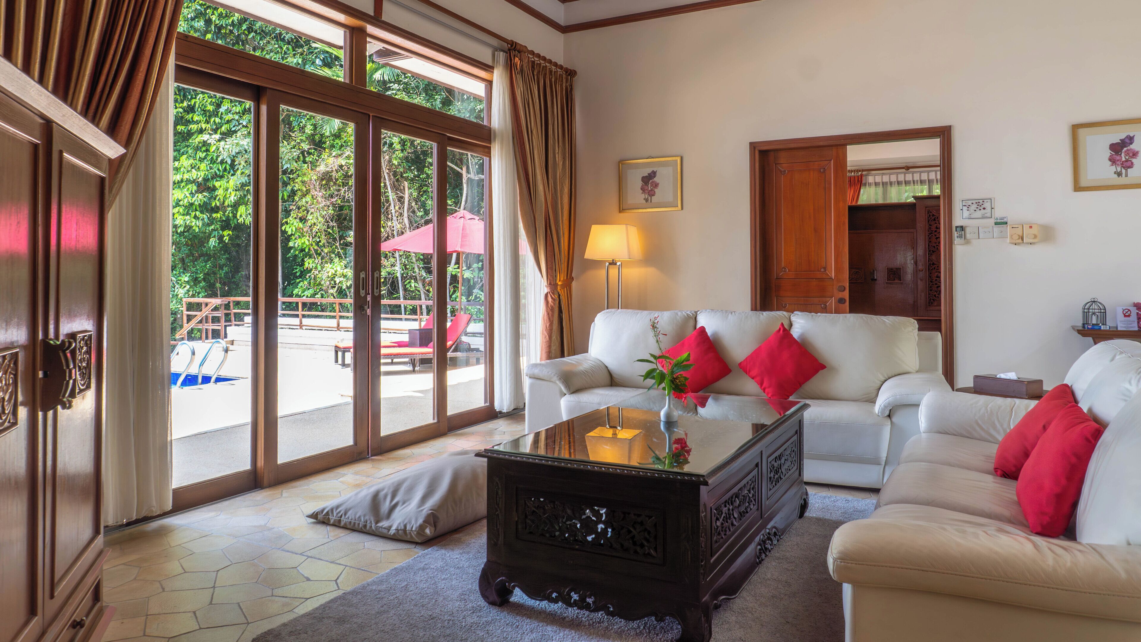 A spacious living room with a white sofa, red cushions, a glass coffee table, and a view of an outdoor pool area.