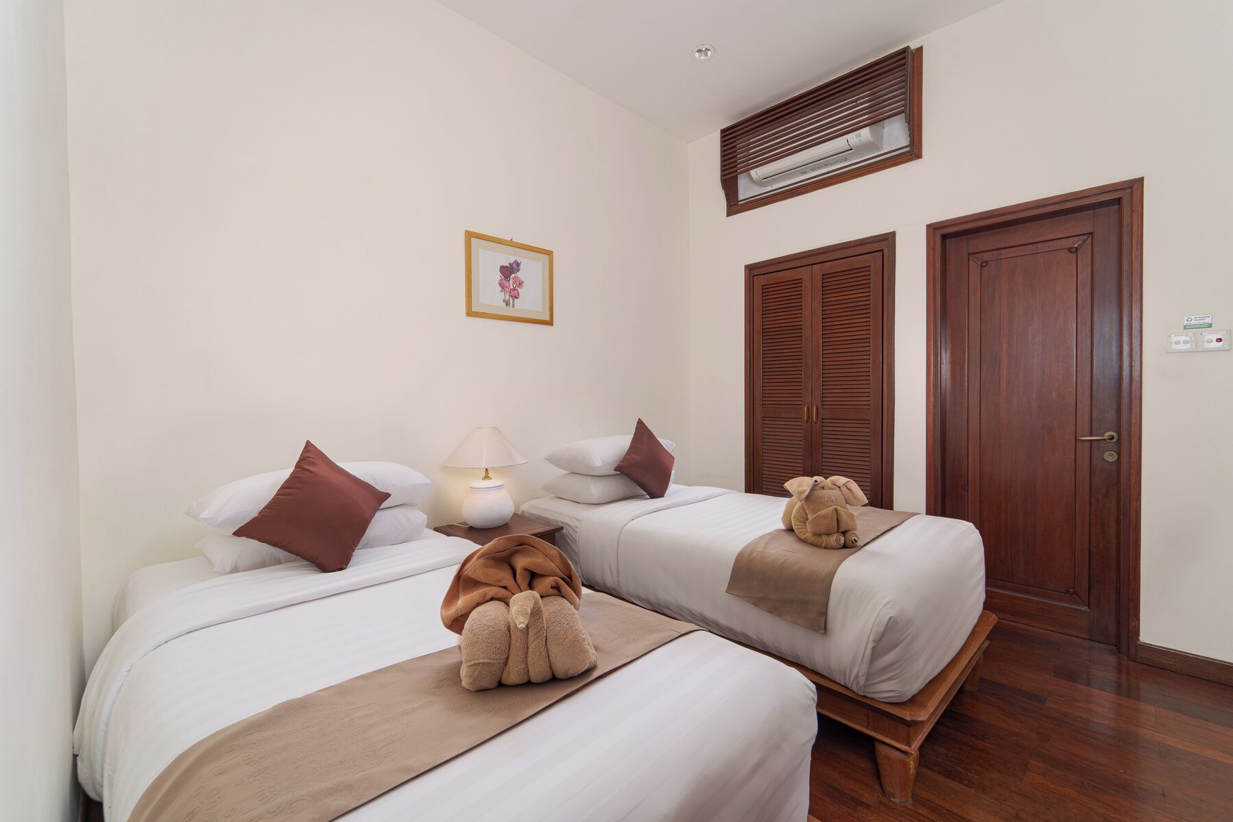 A hotel room with two beds, a bedside lamp, a teddy bear on each bed, a framed picture on the wall, and a wooden wardrobe.