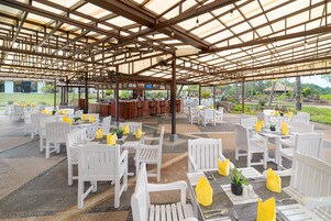 Un área de estar al aire libre, cubierta, con sillas blancas y cojines amarillos, un bar y vistas del paisaje circundante.