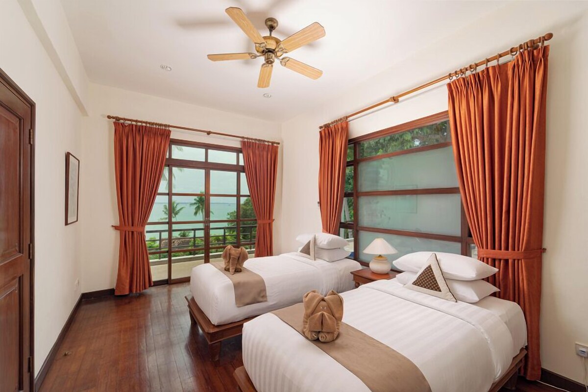 Habitación de hotel con dos camas, ventilador de techo, ventanas amplias con cortinas y vistas al exterior.